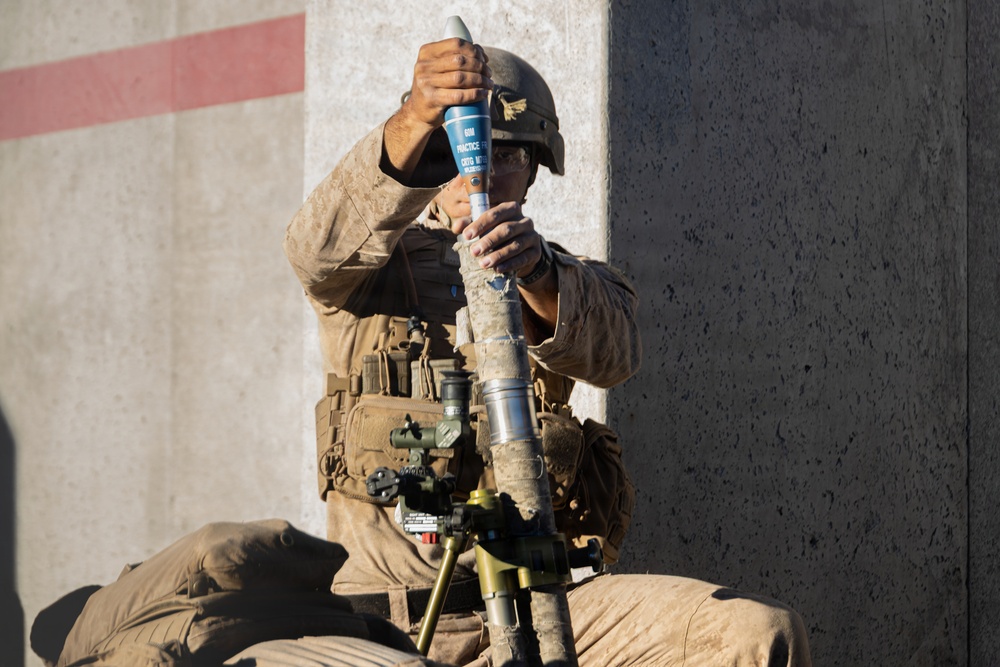 A military service member in desert camouflage uniform and combat helmet is preparing to load a practice mortar round into a mortar tube. The round is visibly marked in blue, indicating it is a training/practice munition. The soldier is kneeling beside a bipod-mounted mortar system against a concrete wall.