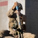 A military service member in desert camouflage uniform and combat helmet is preparing to load a practice mortar round into a mortar tube. The round is visibly marked in blue, indicating it is a training/practice munition. The soldier is kneeling beside a bipod-mounted mortar system against a concrete wall.