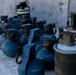 The image shows a collection of blue-painted practice/training grenades arranged on a metal surface. Visible text markings include 'NSN 1330' and 'GRENADE H' along with 'PRACTICE' on at least one unit. These appear to be inert military training devices, identifiable by their blue color coding which is standard for practice/dummy munitions.