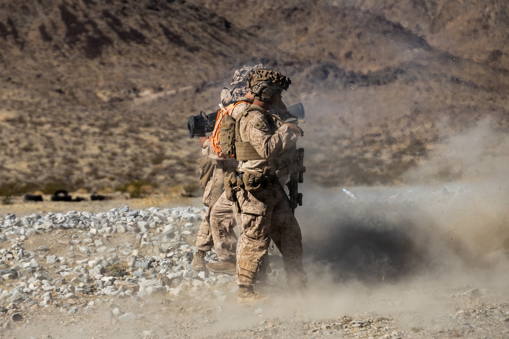 Two military personnel in desert camouflage uniforms are operating what appears to be a shoulder-fired rocket or missile launcher in an arid, rocky desert training environment. A significant dust and smoke cloud is visible to the right, consistent with a weapon discharge or backblast. The setting appears to be a military training range based on the controlled environment and visible range markers in the background.
