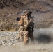 Two military personnel in desert camouflage uniforms are operating what appears to be a shoulder-fired rocket or missile launcher in an arid, rocky desert training environment. A significant dust and smoke cloud is visible to the right, consistent with a weapon discharge or backblast. The setting appears to be a military training range based on the controlled environment and visible range markers in the background.
