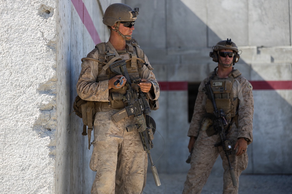 Two armed military personnel in desert camouflage uniforms are positioned near a concrete wall structure. Both are equipped with tactical gear, helmets, body armor, and assault rifles. The setting appears to be an urban or base environment, possibly during a patrol or security operation.