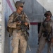 Two armed military personnel in desert camouflage uniforms are positioned near a concrete wall structure. Both are equipped with tactical gear, helmets, body armor, and assault rifles. The setting appears to be an urban or base environment, possibly during a patrol or security operation.
