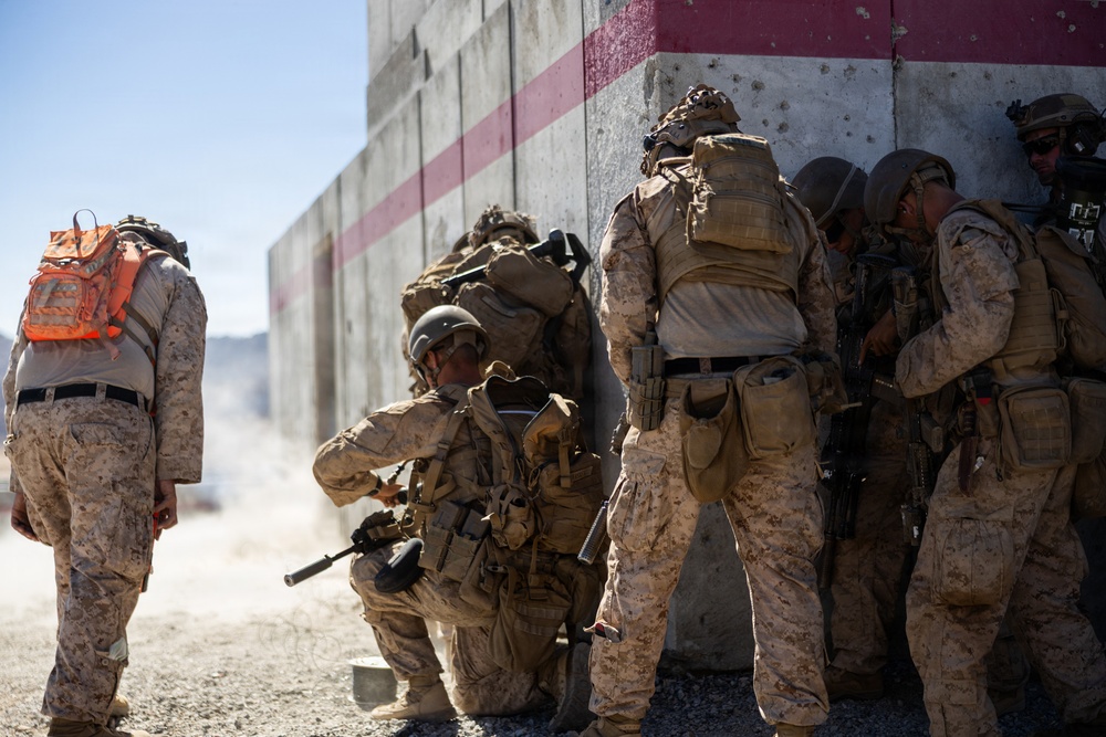 A group of military personnel in desert camouflage uniforms are stacked against a concrete wall with red stripe markings, appearing to conduct a tactical breach or entry exercise. One individual wearing an orange backpack stands apart, likely serving as an observer or safety officer. The setting appears to be a military training facility based on the constructed wall and arid environment.