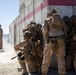 A group of military personnel in desert camouflage uniforms are stacked against a concrete wall with red stripe markings, appearing to conduct a tactical breach or entry exercise. One individual wearing an orange backpack stands apart, likely serving as an observer or safety officer. The setting appears to be a military training facility based on the constructed wall and arid environment.