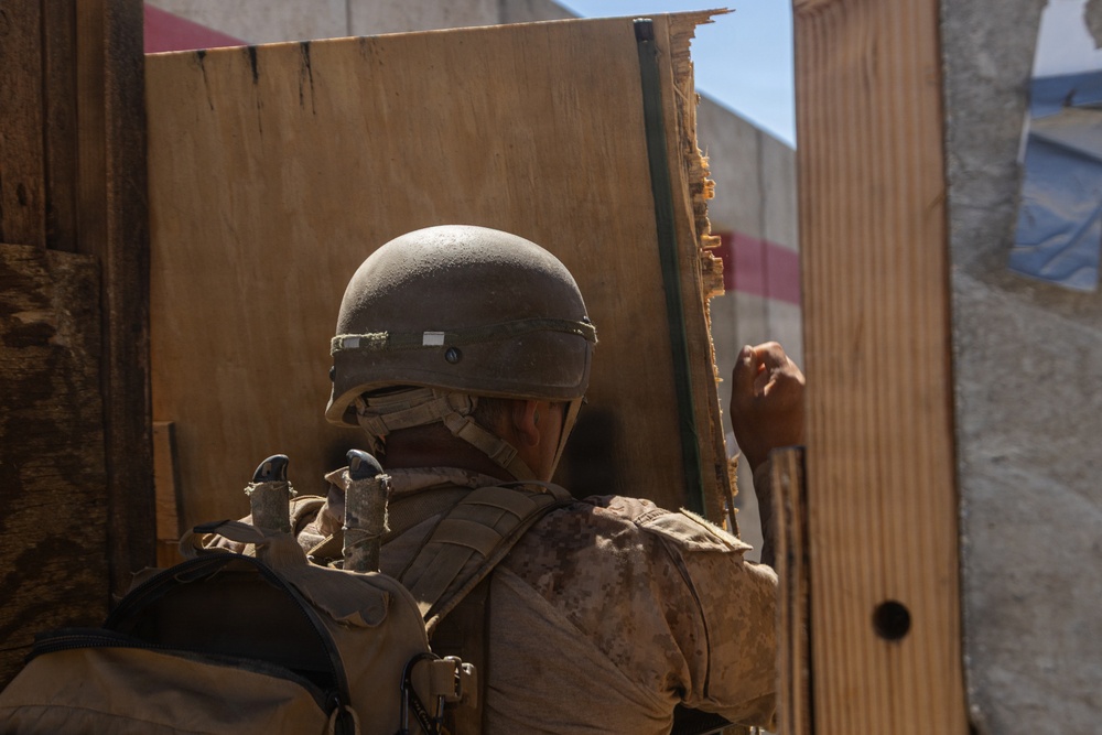 A soldier wearing desert camouflage uniform and a combat helmet is positioned at what appears to be a wooden doorway or barrier. The soldier is viewed from behind and appears to be making a hand signal or knocking on the wooden structure. The setting appears to be an urban or built environment under bright daylight conditions.