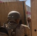 A soldier wearing desert camouflage uniform and a combat helmet is positioned at what appears to be a wooden doorway or barrier. The soldier is viewed from behind and appears to be making a hand signal or knocking on the wooden structure. The setting appears to be an urban or built environment under bright daylight conditions.