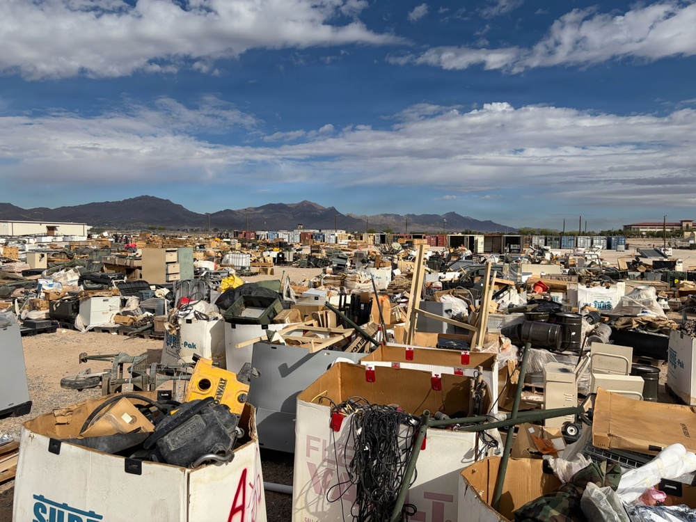 A large outdoor salvage yard or surplus disposal facility filled with an extensive collection of discarded equipment, furniture, tools, and miscellaneous items spread across a wide open area. The scene appears to be located in a desert or arid region, evidenced by the dry terrain, mountain range in the background, and clear southwestern U.S. landscape. The sheer volume and variety of items suggest this may be a military surplus or government disposal yard.