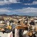 A large outdoor salvage yard or surplus disposal facility filled with an extensive collection of discarded equipment, furniture, tools, and miscellaneous items spread across a wide open area. The scene appears to be located in a desert or arid region, evidenced by the dry terrain, mountain range in the background, and clear southwestern U.S. landscape. The sheer volume and variety of items suggest this may be a military surplus or government disposal yard.