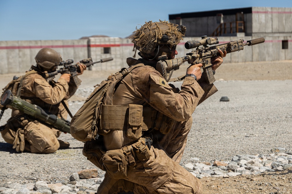 Two military personnel in full tactical gear are positioned on a gravel surface, aiming suppressed rifles toward a concrete structure in the background. Both are wearing desert camouflage uniforms with tactical vests, backpacks, and helmets. The setting appears to be a military training range or exercise area based on the controlled environment and visible range markers.