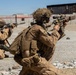 Two military personnel in full tactical gear are positioned on a gravel surface, aiming suppressed rifles toward a concrete structure in the background. Both are wearing desert camouflage uniforms with tactical vests, backpacks, and helmets. The setting appears to be a military training range or exercise area based on the controlled environment and visible range markers.