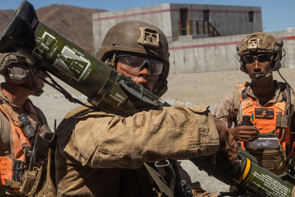 Military personnel are engaged in what appears to be a training exercise in an arid, desert-like environment. The central figure is handling a shoulder-fired rocket launcher (AT4 or similar weapon system) with safety labels visible on the tube. Additional personnel wearing orange safety vests are present, indicating a supervised training or evaluation scenario.