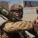 Military personnel are engaged in what appears to be a training exercise in an arid, desert-like environment. The central figure is handling a shoulder-fired rocket launcher (AT4 or similar weapon system) with safety labels visible on the tube. Additional personnel wearing orange safety vests are present, indicating a supervised training or evaluation scenario.
