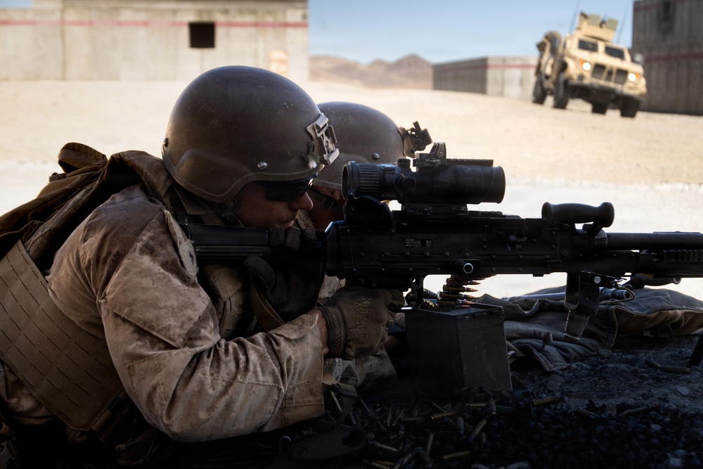 Two military personnel in desert camouflage uniforms are in a prone firing position, with the foreground soldier aiming an M240 machine gun equipped with an optical scope. Spent brass casings are visible on the ground, and an ammunition box is attached to the weapon. A military MRAP vehicle is visible in the background near a building in what appears to be a desert training environment.