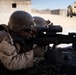 Two military personnel in desert camouflage uniforms are in a prone firing position, with the foreground soldier aiming an M240 machine gun equipped with an optical scope. Spent brass casings are visible on the ground, and an ammunition box is attached to the weapon. A military MRAP vehicle is visible in the background near a building in what appears to be a desert training environment.