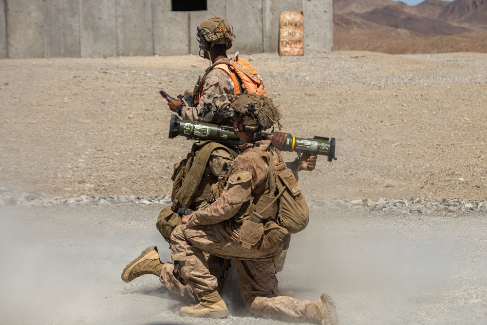 Two military personnel in desert camouflage uniforms are conducting a weapons training exercise at an arid outdoor range. One soldier is kneeling and aiming a rocket launcher (appears to be an AT4 or similar shoulder-fired weapon), while a second standing soldier wearing an orange safety vest observes and monitors. Smoke/backblast is visible near the ground, indicating the weapon has just been or is being fired.