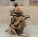 Two military personnel in desert camouflage uniforms are conducting a weapons training exercise at an arid outdoor range. One soldier is kneeling and aiming a rocket launcher (appears to be an AT4 or similar shoulder-fired weapon), while a second standing soldier wearing an orange safety vest observes and monitors. Smoke/backblast is visible near the ground, indicating the weapon has just been or is being fired.