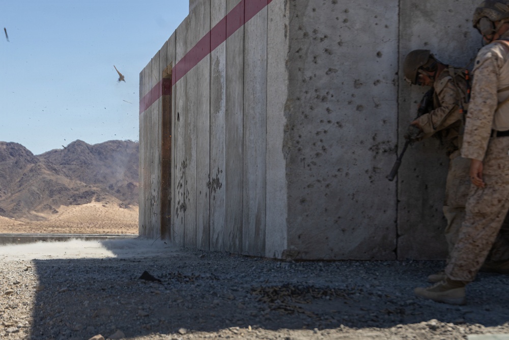 Two military personnel in desert camouflage uniforms and helmets are taking cover behind a concrete wall structure during what appears to be a live-fire or breaching exercise. Debris and fragments are visible in the air to the left of the structure, suggesting a recent explosion or detonation. The setting is an arid desert training environment with rocky mountains visible in the background.