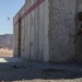 Two military personnel in desert camouflage uniforms and helmets are taking cover behind a concrete wall structure during what appears to be a live-fire or breaching exercise. Debris and fragments are visible in the air to the left of the structure, suggesting a recent explosion or detonation. The setting is an arid desert training environment with rocky mountains visible in the background.
