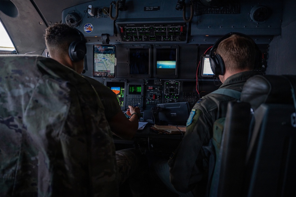 Two military aircrew members are seated in the cockpit of a large military transport aircraft, viewed from behind. Both personnel are wearing flight suits and aviation headsets while operating the aircraft's flight systems. Multiple digital displays, navigation screens, and instrument panels are visible throughout the cockpit.