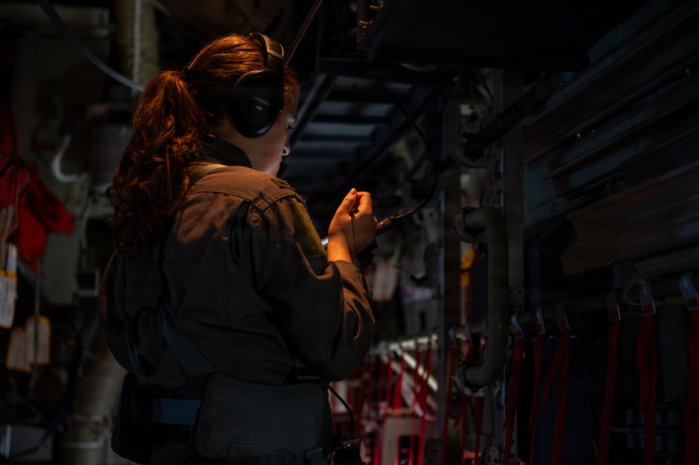 A female military personnel member wearing a flight suit and aviation headset is working inside what appears to be a military cargo aircraft. She is illuminated by a small handheld light while performing an inspection or maintenance task on the aircraft's interior wall systems. The environment is dark with visible cables, straps, and mechanical components along the fuselage wall.