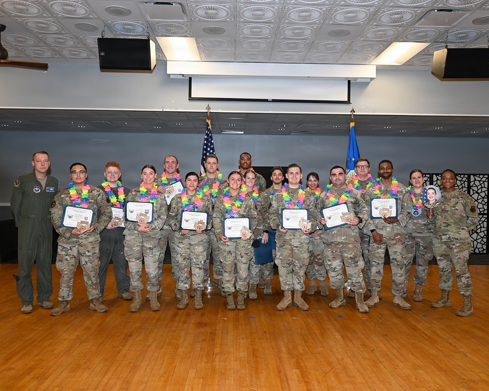 A group of approximately 16 military personnel in camouflage uniforms pose for a formal group photograph in what appears to be a base auditorium or event hall. Most of the recipients are wearing colorful Hawaiian leis and holding certificates and wooden plaques, indicating a recognition or award ceremony. Two individuals on the far left and far right appear to be presiding officers not wearing leis.