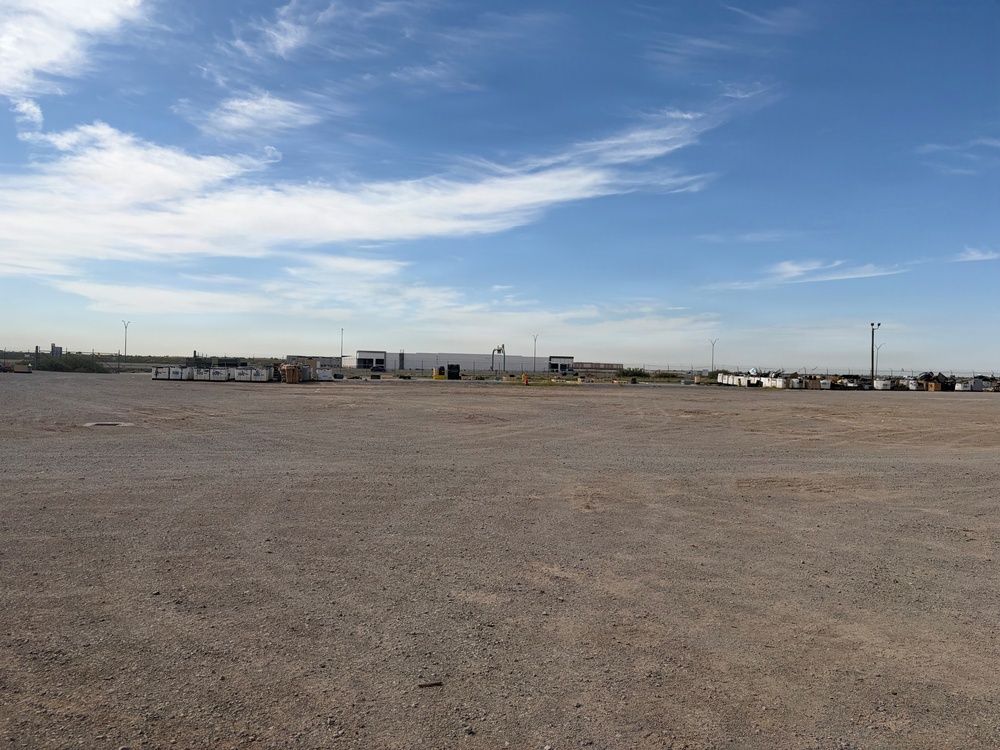 A large, flat gravel or dirt yard typical of an industrial or oilfield services facility, photographed under a partly cloudy blue sky. The foreground is dominated by an expansive unpaved surface, while the background shows a row of white storage containers, a large warehouse or industrial building, utility poles, and miscellaneous equipment. The scene suggests an active staging or laydown yard in an arid region.