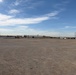 A large, flat gravel or dirt yard typical of an industrial or oilfield services facility, photographed under a partly cloudy blue sky. The foreground is dominated by an expansive unpaved surface, while the background shows a row of white storage containers, a large warehouse or industrial building, utility poles, and miscellaneous equipment. The scene suggests an active staging or laydown yard in an arid region.