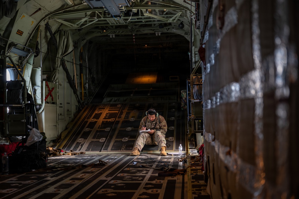A military service member in camouflage uniform sits on the cargo floor of a large military transport aircraft, writing on a clipboard or notepad. The aircraft's interior shows a wide cargo bay with roller-track flooring, structural ribs, and secured cargo pallets visible to the right. The lighting is dim with a warm ambient glow from the rear ramp area.