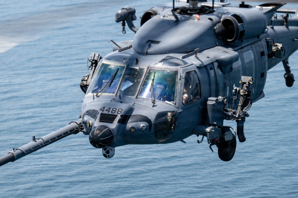 A military helicopter, marked with the tail number 4488, is photographed in close-up while flying low over open water. The aircraft appears to be a Sikorsky HH-60 Pave Hawk or similar variant, painted in a dark gray military finish. The image captures fine detail of the cockpit, sensors, refueling probe, and crew members visible through the canopy.