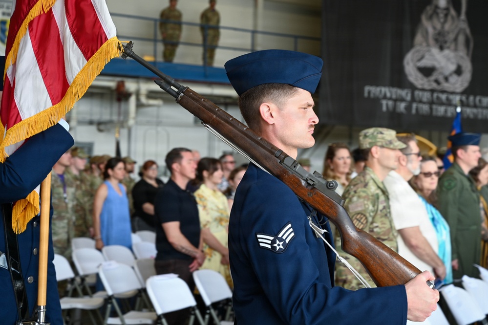 A U.S. Air Force color guard ceremony is taking place inside what appears to be a military hangar. A Senior Airman in dress blues carries an M1 Garand rifle at shoulder arms while a U.S. flag with gold fringe is visible to the left. An audience of military personnel and civilians stands in attendance behind white folding chairs.
