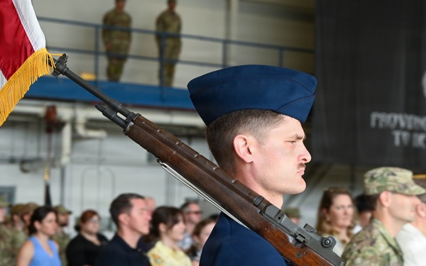 A U.S. Air Force color guard ceremony is taking place inside what appears to be a military hangar. A Senior Airman in dress blues carries an M1 Garand rifle at shoulder arms while a U.S. flag with gold fringe is visible to the left. An audience of military personnel and civilians stands in attendance behind white folding chairs.