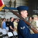 A U.S. Air Force color guard ceremony is taking place inside what appears to be a military hangar. A Senior Airman in dress blues carries an M1 Garand rifle at shoulder arms while a U.S. flag with gold fringe is visible to the left. An audience of military personnel and civilians stands in attendance behind white folding chairs.
