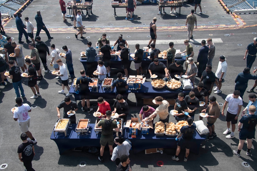 An aerial view of a large outdoor buffet-style meal event taking place on what appears to be the flight deck of a naval vessel. Dozens of personnel in casual and military attire are lined up along blue-draped serving tables loaded with food. The event resembles a morale or holiday cookout commonly held aboard U.S. Navy ships.