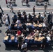 An aerial view of a large outdoor buffet-style meal event taking place on what appears to be the flight deck of a naval vessel. Dozens of personnel in casual and military attire are lined up along blue-draped serving tables loaded with food. The event resembles a morale or holiday cookout commonly held aboard U.S. Navy ships.