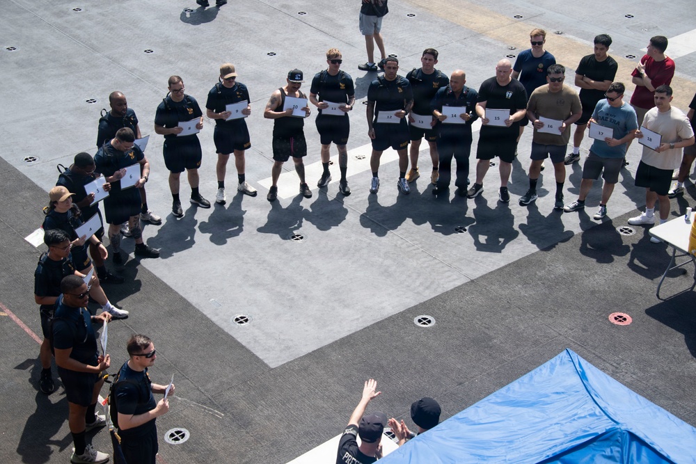 A group of military personnel, likely U.S. Navy sailors, are assembled on what appears to be the flight deck of an aircraft carrier. Participants are holding numbered placards ranging from approximately 4 to 18, suggesting a competition or organized event. An official or instructor at the bottom of the frame appears to be addressing or directing the group.