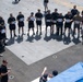 A group of military personnel, likely U.S. Navy sailors, are assembled on what appears to be the flight deck of an aircraft carrier. Participants are holding numbered placards ranging from approximately 4 to 18, suggesting a competition or organized event. An official or instructor at the bottom of the frame appears to be addressing or directing the group.