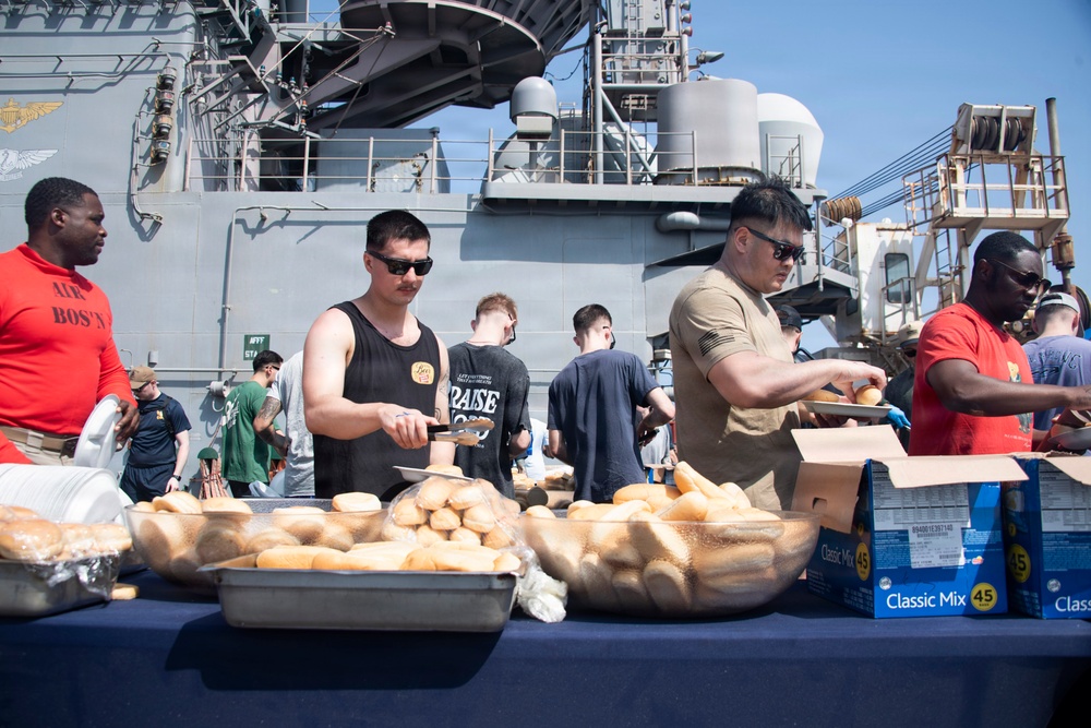 A group of people are serving food at an outdoor cookout or barbecue event on the deck of a naval vessel. Multiple individuals are handling buns and food items at a table covered with trays of bread rolls and boxed supplies. The setting appears to be a military ship, likely during a morale or recreational event for crew members.