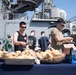 A group of people are serving food at an outdoor cookout or barbecue event on the deck of a naval vessel. Multiple individuals are handling buns and food items at a table covered with trays of bread rolls and boxed supplies. The setting appears to be a military ship, likely during a morale or recreational event for crew members.