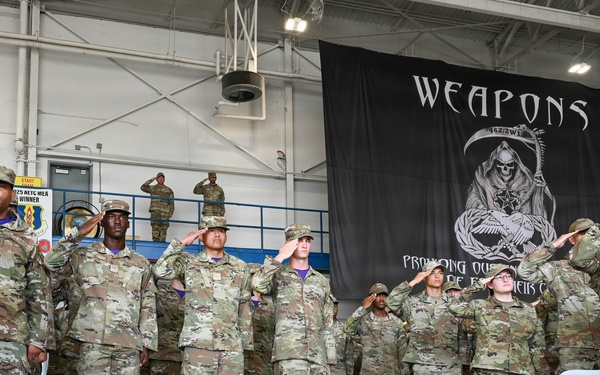 A military formation ceremony is taking place inside a hangar facility, with personnel in OCP (Operational Camouflage Pattern) uniforms rendering salutes. A guidon bearer on the left holds a blue flag marked '33 MXS' (33rd Maintenance Squadron). A large black banner reading 'WEAPONS' with a grim reaper graphic is prominently displayed in the background.