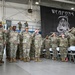 A military formation ceremony is taking place inside a hangar facility, with personnel in OCP (Operational Camouflage Pattern) uniforms rendering salutes. A guidon bearer on the left holds a blue flag marked '33 MXS' (33rd Maintenance Squadron). A large black banner reading 'WEAPONS' with a grim reaper graphic is prominently displayed in the background.