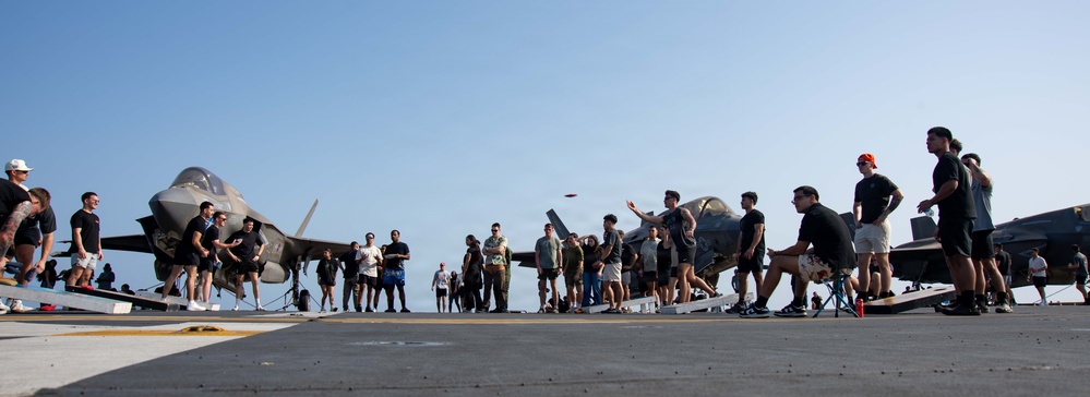 A group of military personnel are gathered on what appears to be an aircraft carrier flight deck, engaging in recreational activities. Multiple F-35 stealth fighter jets are parked in the background on the deck. The scene suggests a morale or leisure event during a deployment.