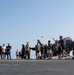 A group of military personnel are gathered on what appears to be an aircraft carrier flight deck, engaging in recreational activities. Multiple F-35 stealth fighter jets are parked in the background on the deck. The scene suggests a morale or leisure event during a deployment.