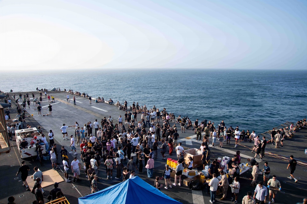 A large gathering of people is taking place on the flight deck of a naval vessel at sea. The event appears to be an informal social gathering or morale event, with food and beverage tables set up and personnel in casual civilian clothing mingling freely. The ship's deck extends toward the bow with the open ocean visible on multiple sides.