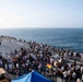 A large gathering of people is taking place on the flight deck of a naval vessel at sea. The event appears to be an informal social gathering or morale event, with food and beverage tables set up and personnel in casual civilian clothing mingling freely. The ship's deck extends toward the bow with the open ocean visible on multiple sides.