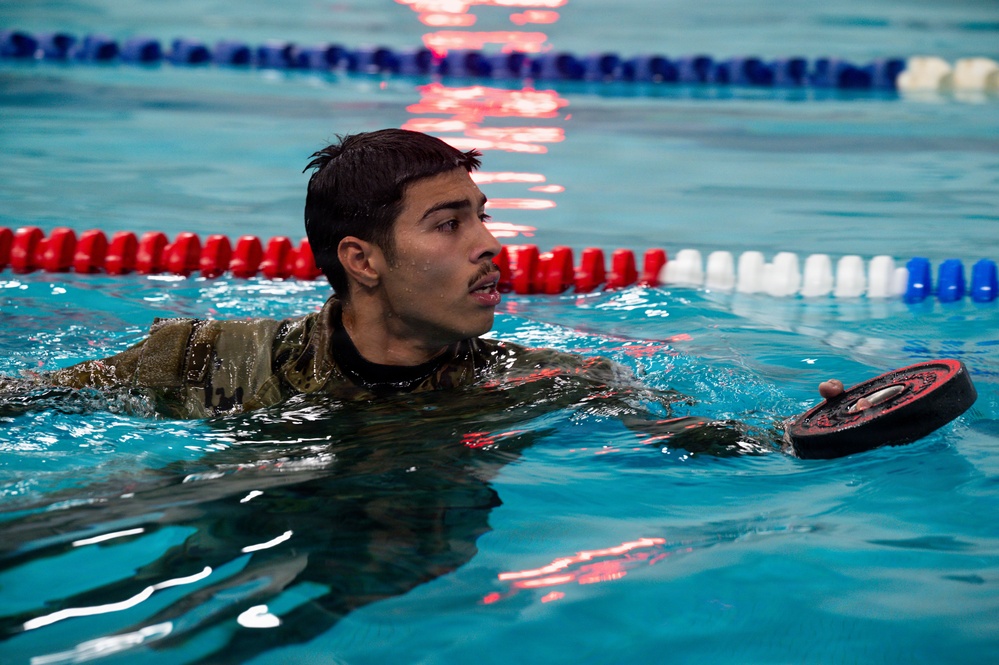 A young male in military camouflage uniform is swimming in an indoor pool while holding a weighted barbell plate. The individual appears to be participating in a military fitness or water survival training exercise. The pool features standard lane dividers in red, white, and blue colors.
