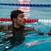 A young male in military camouflage uniform is swimming in an indoor pool while holding a weighted barbell plate. The individual appears to be participating in a military fitness or water survival training exercise. The pool features standard lane dividers in red, white, and blue colors.