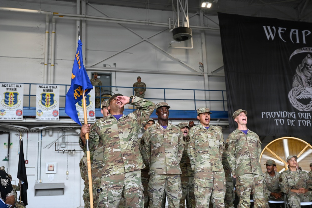 A group of U.S. Air Force personnel in OCP (Operational Camouflage Pattern) uniforms are shouting or cheering enthusiastically during what appears to be a military ceremony or competition event inside an aircraft hangar. One airman in the foreground is holding a blue unit guidon flag while calling out loudly. Banners in the background indicate '2023 AETC MEA Winner' and '2024 AETC MEA Winner' awards.