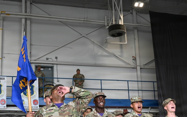 A group of U.S. Air Force personnel in OCP (Operational Camouflage Pattern) uniforms are shouting or cheering enthusiastically during what appears to be a military ceremony or competition event inside an aircraft hangar. One airman in the foreground is holding a blue unit guidon flag while calling out loudly. Banners in the background indicate '2023 AETC MEA Winner' and '2024 AETC MEA Winner' awards.