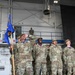 A group of U.S. Air Force personnel in OCP (Operational Camouflage Pattern) uniforms are shouting or cheering enthusiastically during what appears to be a military ceremony or competition event inside an aircraft hangar. One airman in the foreground is holding a blue unit guidon flag while calling out loudly. Banners in the background indicate '2023 AETC MEA Winner' and '2024 AETC MEA Winner' awards.